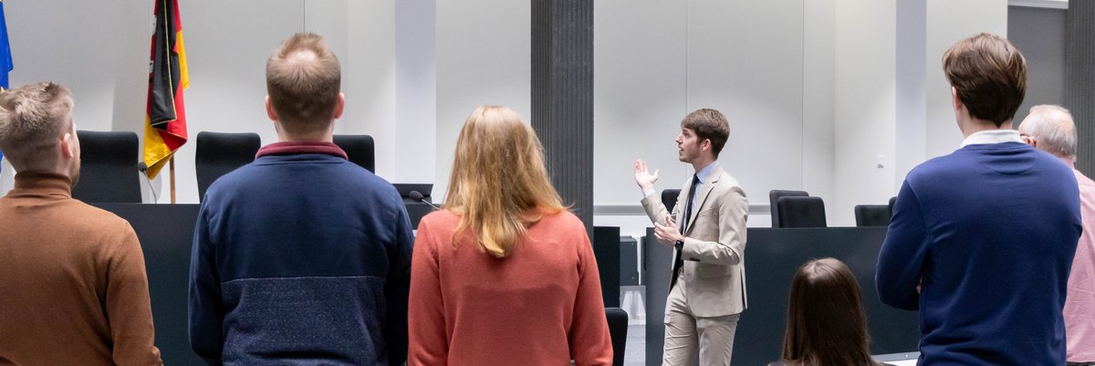 Mehrere Besucherinnen und Besucher stehen mit dem Rücken zur Kamera und sehen sich im Forum des Niedersächsischen Landtages um, während ein Landtagslotse neben dem Redepult steht und in den Raum deutet.