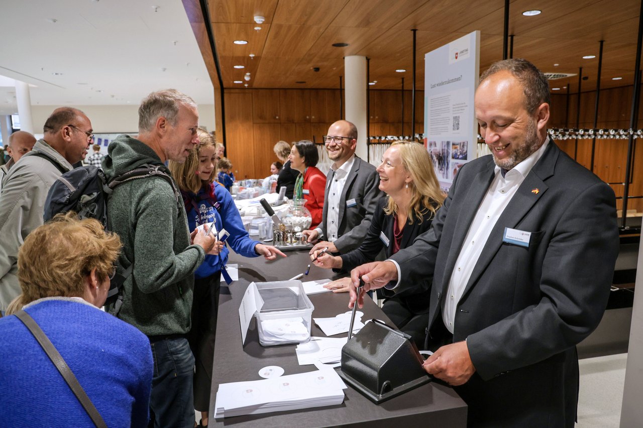 Links einige Besucherinnen und Besucher, ihnen gegenüber drei lachende Mitarbeitende des Stenografischen Dienstes mit Button-Maschine, Notizblock und Stanze.