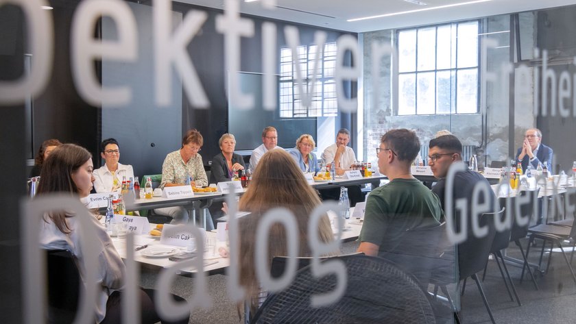 Blick durch eine Glasscheibe in einen Raum mit einem großen Konferenztisch. Daran sitzen die Mitglieder des Landtagspräsidiums und gegenüber einige Auszubildende der Salzgitter Flachstahl AG. Die Glasscheibe ist beschriftet; unter anderem ist das Wort "Dialog" zu lesen.