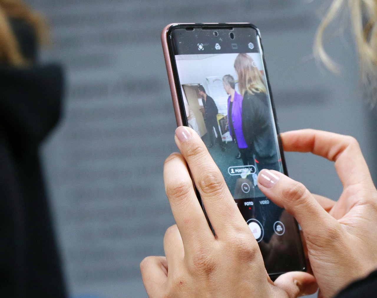 Im Vordergrund ein Smartphone mit geöffneter Kamera-App in den Händen einer jungen Frau. Im Hintergrund die Landtagspräsidenten im Austausch mit Ehrenamtlichen.