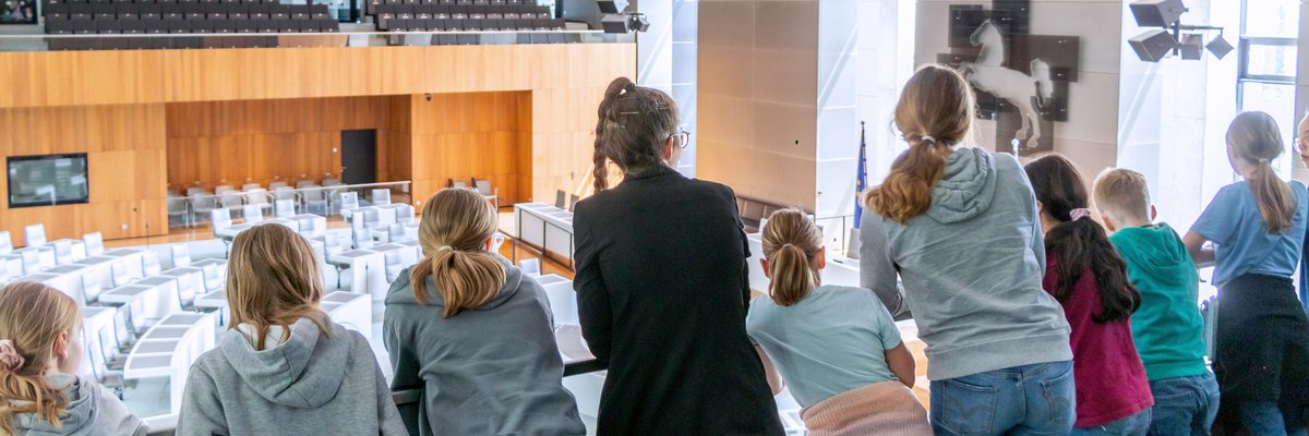 Bild: © Niedersächsischer Landtag Eine Landtagslotsin umringt von Kindern schaut mit ihnen von der Besuchertribüne hinunter in den leeren Plenarsaal.