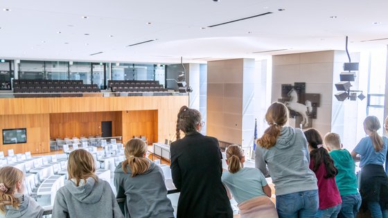 Bild: © Niedersächsischer Landtag Eine Landtagslotsin umringt von Kindern schaut mit ihnen von der Besuchertribüne hinunter in den leeren Plenarsaal.