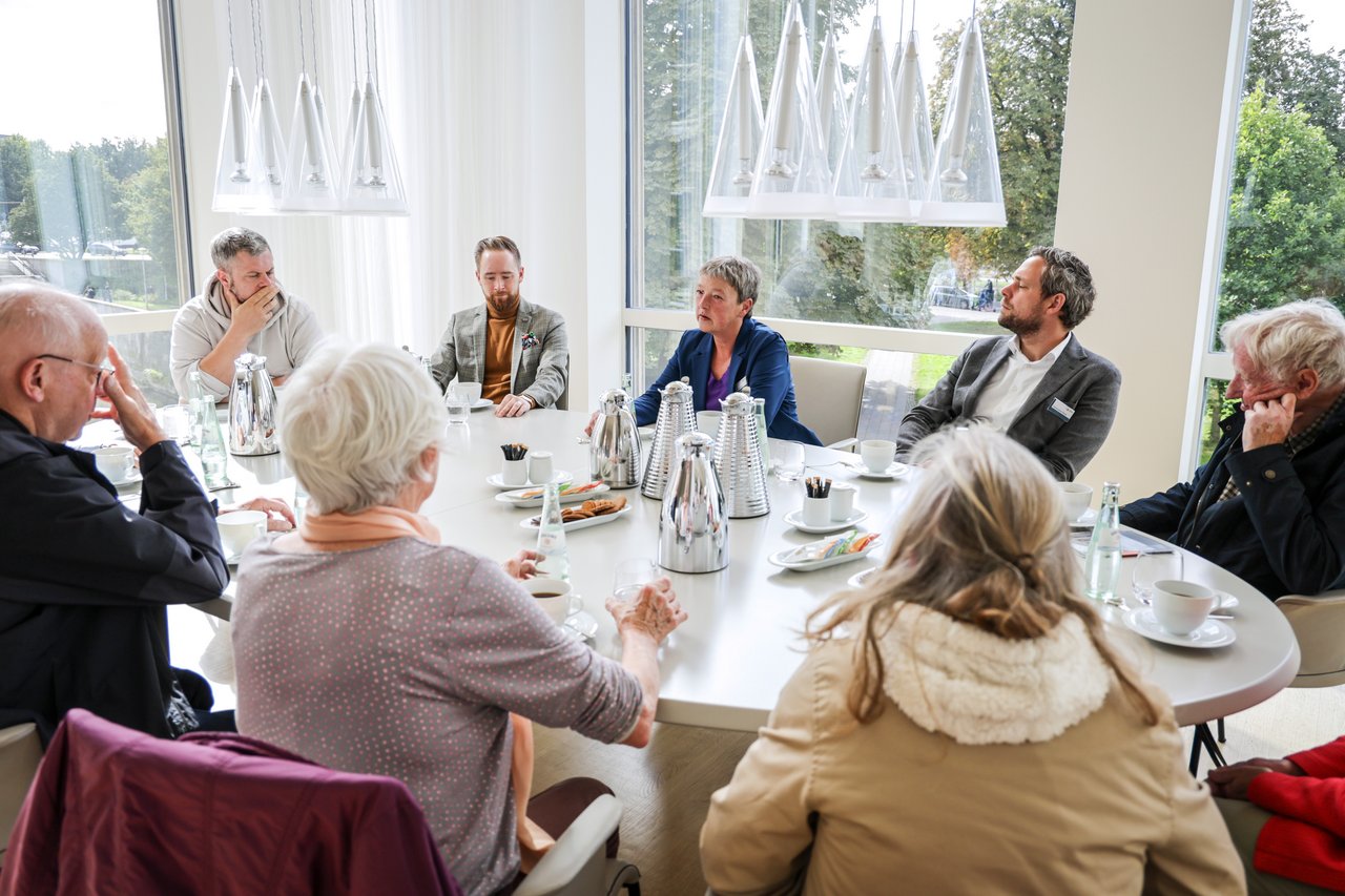 Landtagspräsidentin Hanna Naber sitzt inmitten einer kleinen Gruppe von Besucherinnen und Besuchern am Konferenztisch in ihrem Arbeitszimmer. Auf dem Tisch stehen Kannen, Tassen und Teller mit Keksen.