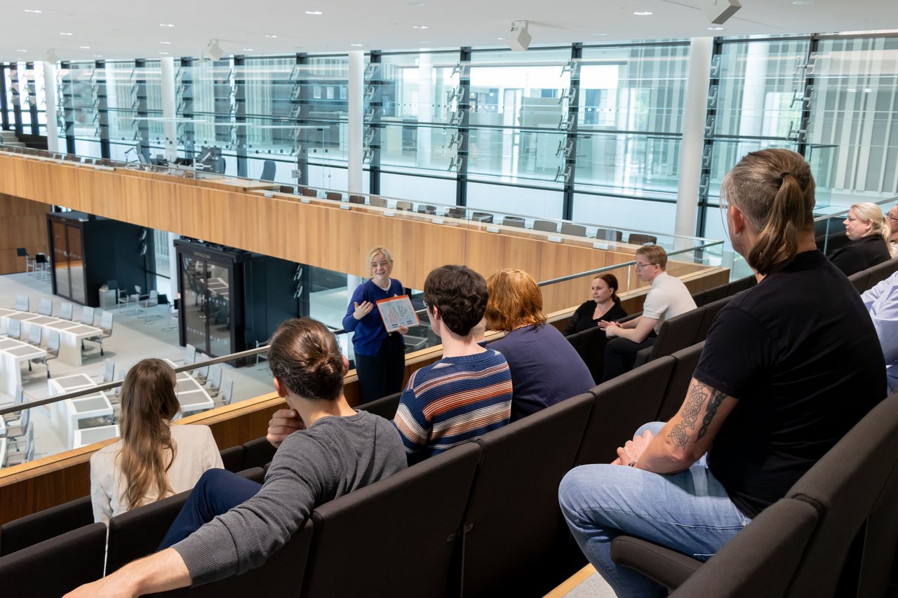 Landtagslotsin Laura Koren steht vor einer Gruppe Menschen auf der Besuchertribüne im Plenarsaal und erklärt etwas anhand eines Schaubildes