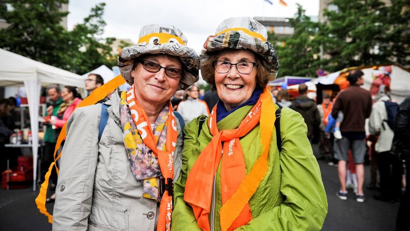 Zwei Damen mit bunten Hüten aus Zeitungen und orangen Schals lachen in die Kamera beim Kirchentag in Berlin 2017 (Abend der Begegnung)